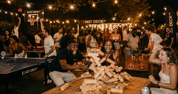 A Hot Deck setup at night — giant Jenga collapsing across the table, with table tennis, beer pong, ring toss and basketball under string lights